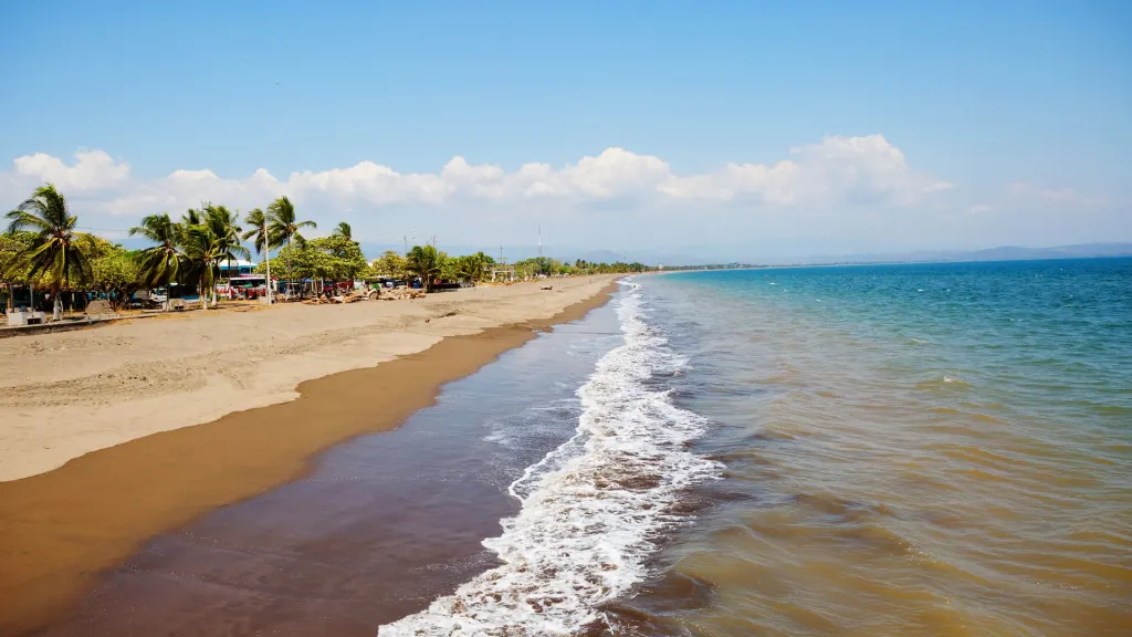 A wide sandy beach with gentle waves under a clear blue sky. Palm trees and small beach structures line the shore on the left.
